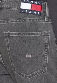 Grå denimjeans med en distinkt bakficka som har en liten Tommy Jeans-logotyp i rött, vitt och bl ått. Sömmen är synlig.