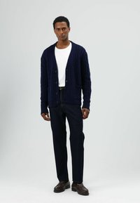 Marineblauer Strickcardigan mit Knöpfen, getragen über einem weißen Hemd. Kombiniert mit dunklen Jeans und braunen Schuhen. Verfügt über Vordertaschen und gerippte Bündchen.