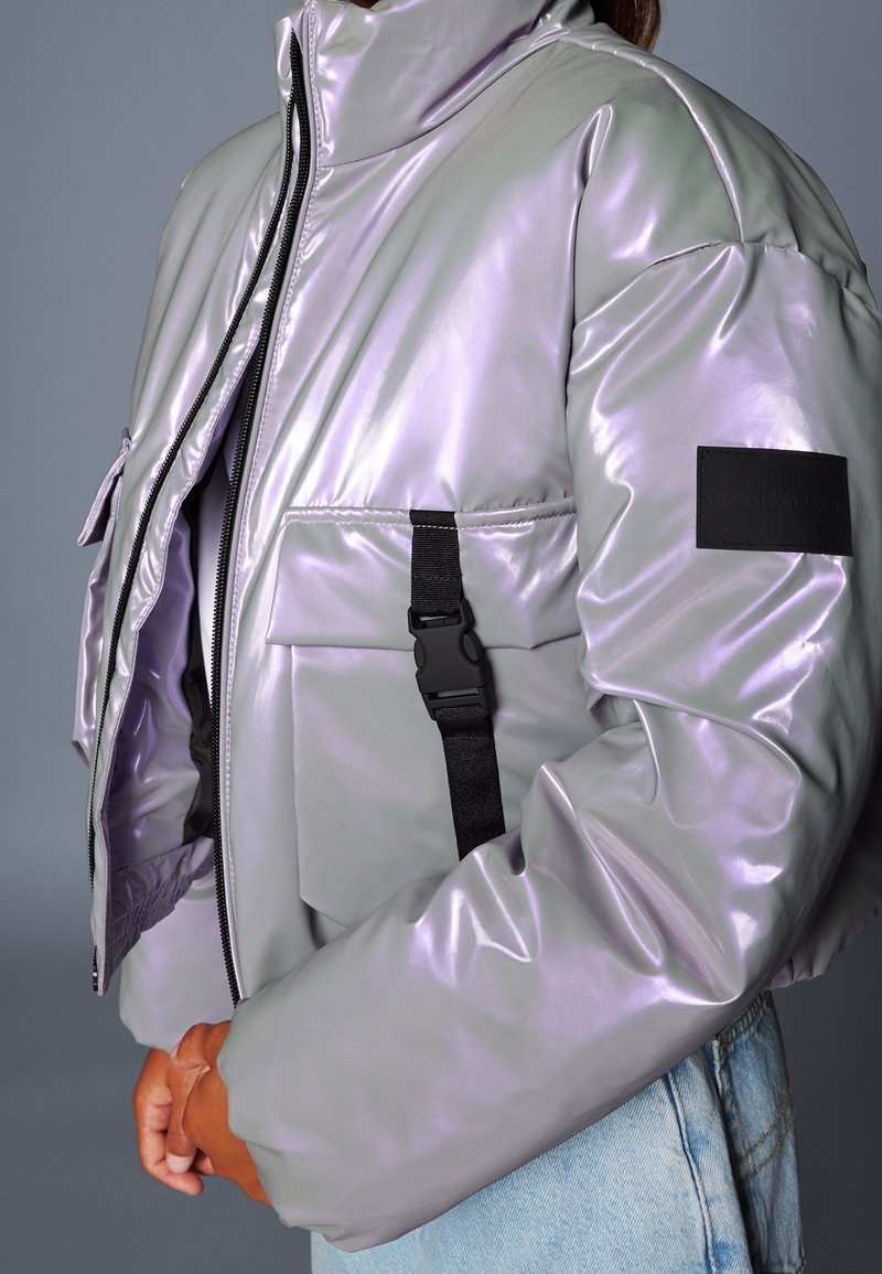 Osoba ubrana w błyszczącą, jasnofioletową kurtkę puchową z czarnym paskiem z klamrą na przedniej kieszeni oraz jasno-niebieskie dżinsy.