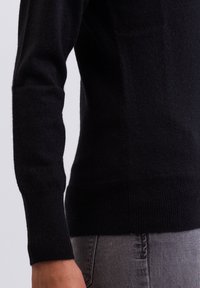 Svart ulltröja med en åtsittande design, ribbade muddarna och kanten, samt en mjuk textur. Bilden fokuserar på sidovyn och ärmdetaljerna.