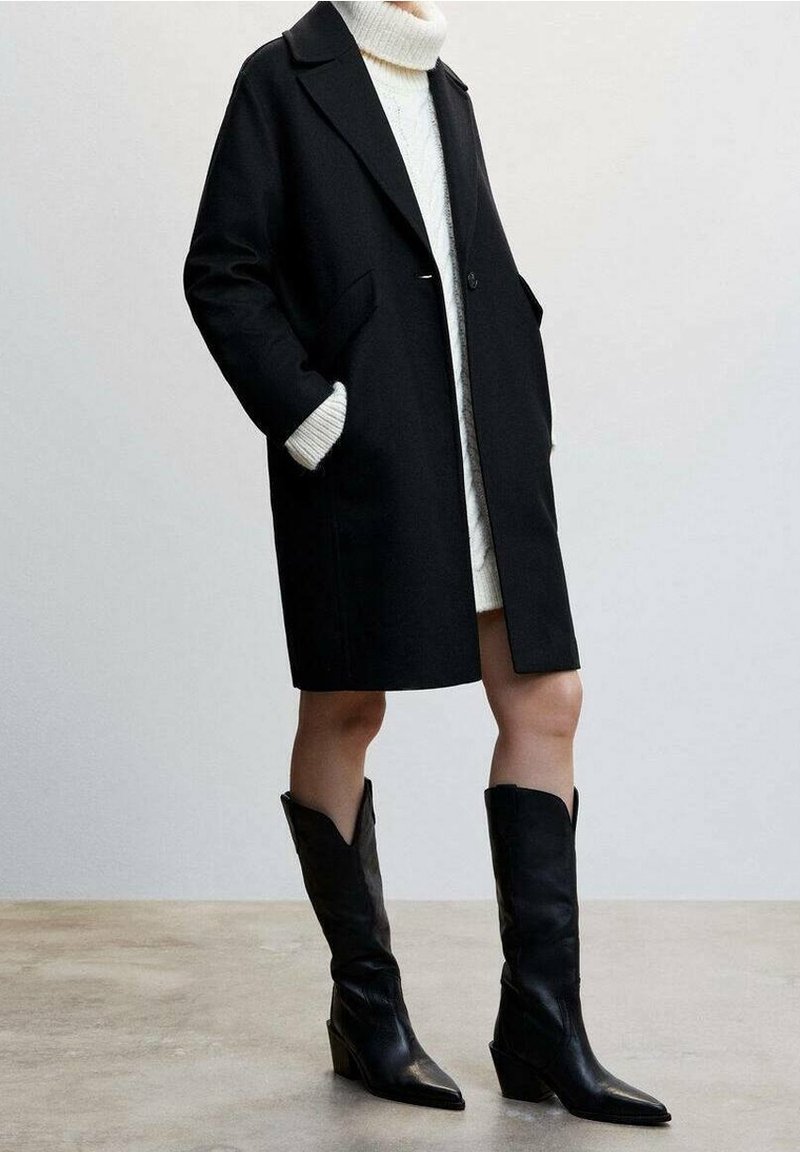 Personne portant un manteau noir mi-long au genou sur une robe-pull à col roulé blanche et des bottes noires à bout pointu mi-mollet, debout sur un sol uni.