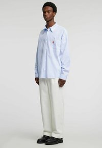 Stripet langermet skjorte i lyseblått og hvitt, med en enkelt brystlomme med en liten logo, kombinert med kremfargede vidbente bukser og svarte sko.