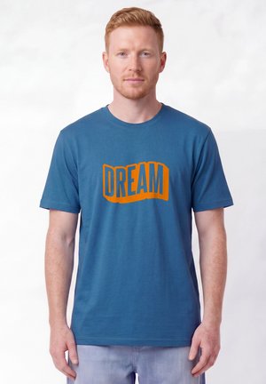 Mann mit heller Haut und roten Haaren, der ein blaues T-Shirt mit orangefarbenem "DREAM"-Text trägt und vor einem schlichten hellen Hintergrund steht.