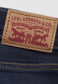 Tumman denim-kankaan päällä on tan-värinen nahkalaatta, jossa on punaisia tekstejä ja kuvastoja; siinä on hevonen, cowboyt ja farkut, ja se on ommeltu kontrastoivalla keltaisella langalla.