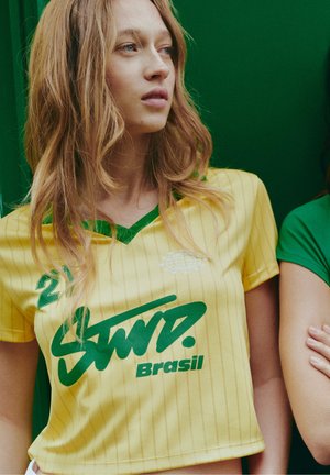 Femeie cu păr lung blond, purtând un tricou sport galben al Braziliei cu litere verzi, stând în fața unui fundal verde.