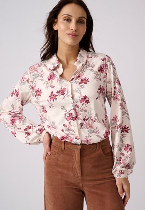 Frau trägt eine langärmelige weiße Bluse mit pinkem Blumenmuster und braune Cordhosen, posiert mit einer Hand in der Hüfte vor einfachem Hintergrund.