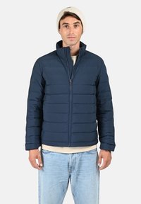 Doudoune bleu marine avec un design matelassé horizontal, col montant, fermeture éclair à l'avant et poignets légèrement élastiques. Le modèle porte un jean clair et un bonnet léger.