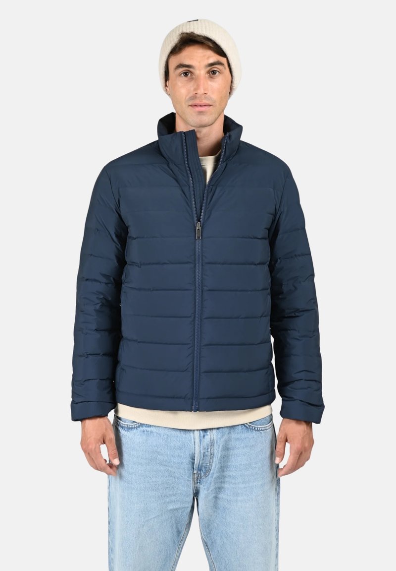 Doudoune bleu marine avec un design matelassé horizontal, col montant, fermeture éclair à l'avant et poignets légèrement élastiques. Le modèle porte un jean clair et un bonnet léger.