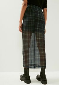 Jupe en plaid transparent vert foncé et noir à coupe droite, dotée d'une fente au dos et d'une texture brillante. Associée à des bottines noires.