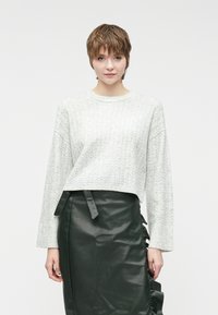 Ανοιχτό γκρι πλεκτό πουλόβερ με χαμηλούς ώμους και φαρδιά μανίκια, συνδυασμένο με μια σκούρα πράσινη δερμάτινη φούστα που διαθέτει πλαϊνές ρικτές και κορδόνια.