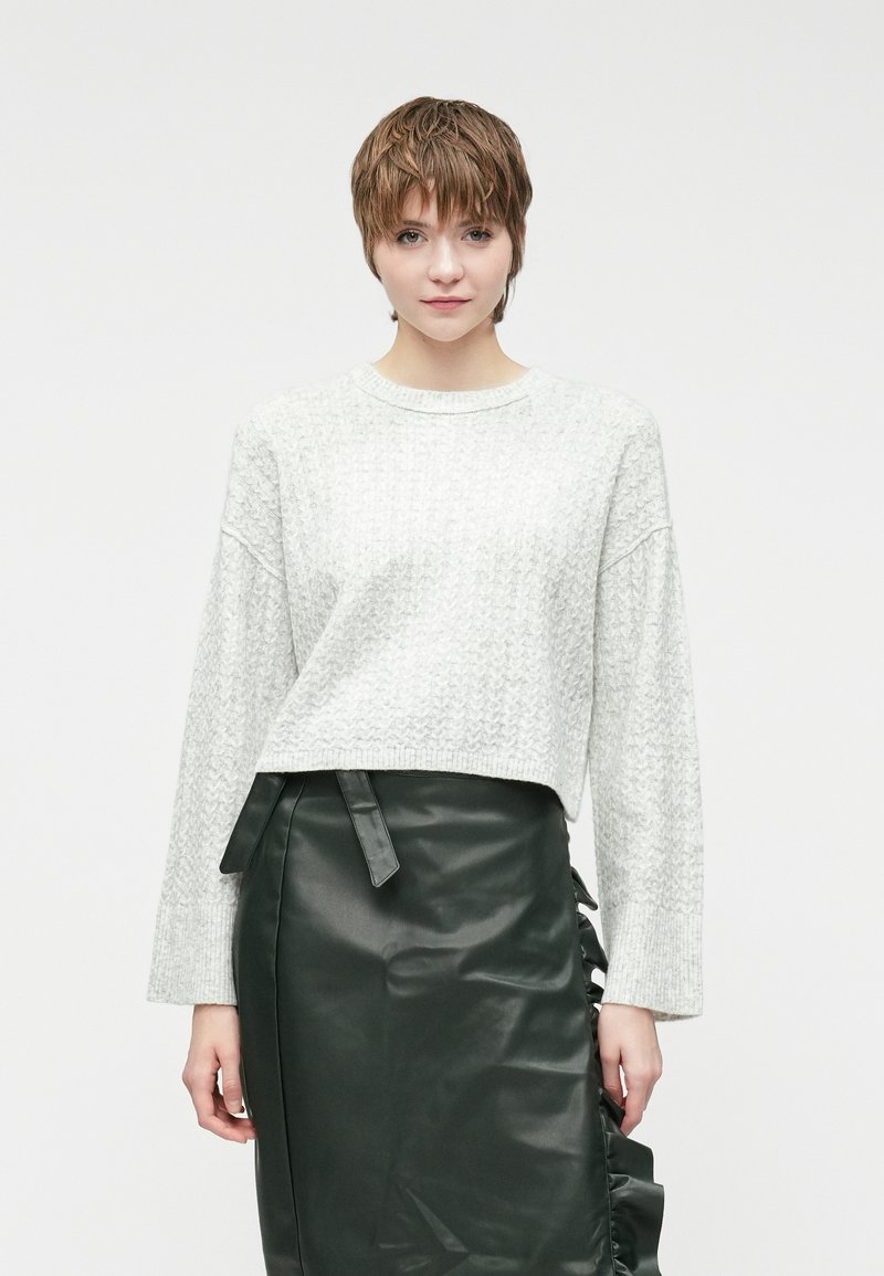 Ανοιχτό γκρι πλεκτό πουλόβερ με χαμηλούς ώμους και φαρδιά μανίκια, συνδυασμένο με μια σκούρα πράσινη δερμάτινη φούστα που διαθέτει πλαϊνές ρικτές και κορδόνια.