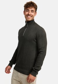 Mørkegrønn kvart-zip genser med et teksturert herringbone-mønster, ribbestrikkede mansjetter og krage. Laget av et mykt, strikket stoff.