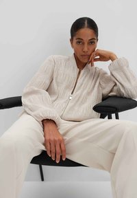 Hellbeige Bomberjacke mit strukturiertem vertikalem Nähten, Reißverschluss und gerafften Bündchen, getragen über cremefarbenen Hosen. Auf einem neutralen Hintergrund.