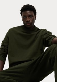 Felpa girocollo verde scuro con polsini e orlo a costa. Texture liscia e vestibilità rilassata, indossata con pantaloni abbinati. Design semplice e minimalista.