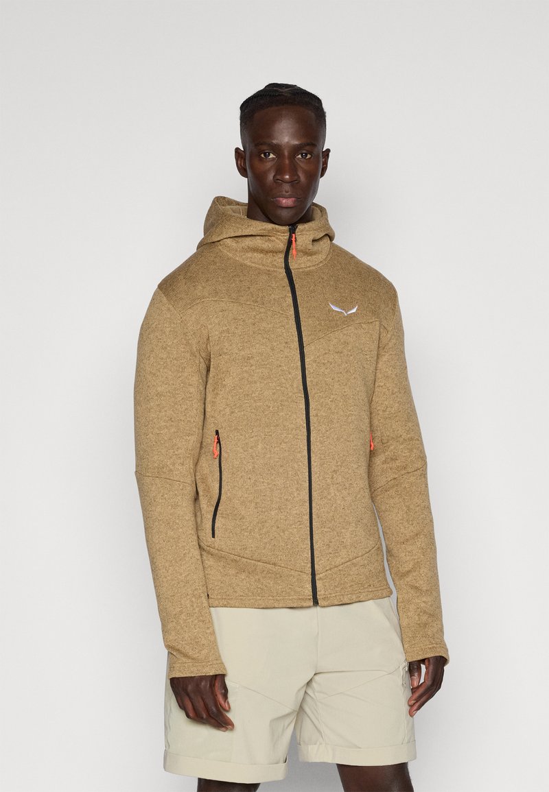 Brūns zip-up kapuce izgatavota no strukturēta materiāla ar pievienotu kapuci, melnu rāvējslēdzēju un oranžiem akcentiem. Iekļauj sānu kabatas un ģeometriskas šuves.