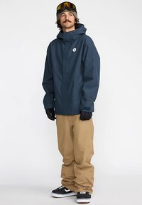 Veste imperméable bleu marine avec capuche, pantalon de neige beige, gants noirs et baskets noires avec accents blancs. Présente un logo sur la poitrine.