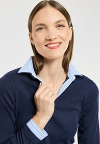 Donna sorridente con capelli castani lisci che indossa una maglia blu navy sopra una camicia a righe azzurra con colletto, mentre tiene il colletto.