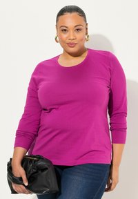 Fuchsia Langarm-T-Shirt mit Rundhalsausschnitt, kombiniert mit dunkelblauen Jeans, gehalten von einer schwarzen Handtasche. Glatte Textur, figurnahe Passform.