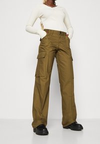 Cargo kalhoty z olivově zelené látky s širokým střihem, s několika bočními kapsami a zapínáním na knoflík. Oblékáno s bílým topem s dlouhým rukávem.