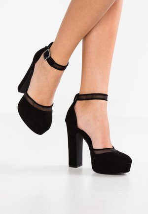 Piernas que llevan plataformas de tacón alto de ante negro con correas en los tobillos, posicionadas con un pie ligeramente elevado sobre un fondo blanco.