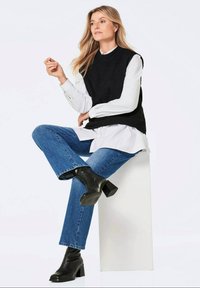 Schwarze gestrickte ärmellose Weste über einem weißen Button-Up-Shirt, kombiniert mit blauen geraden Jeans und schwarzen Stiefeletten mit Absatz.