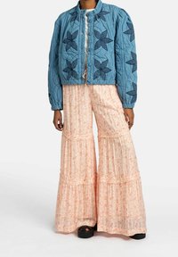 Veste bomber matelassée bleue avec un motif floral, associée à un pantalon léger rose pâle à volants et imprimé floral, portée avec des chaussures noires.