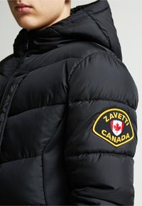 Melna pūka jaka ar kapuci, teksturēta auduma, un uz piedurknes esošu uzlīmi ar "Zavetti Canada" un kļavas lapas emblemu.