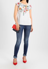 Blusa blanca con bordados florales coloridos, mangas capadas enrolladas, combinada con jeans azul oscuro y zapatos de tacón rojo. Bolso negro y rojo.