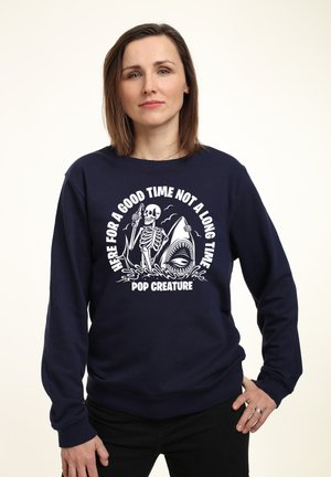Mørkeblå sweatshirt med et grafisk tryk af et skelet med en haj og bølger, hvid tekst med ordene "HER FOR AT HAVE EN GOD TID, IKKE EN LANG TID."