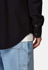 Sort langærmet skjorte med knapmanchetter, båret over en hvid t-shirt. Lys blå jeans med en subtil tekstur og detaljer ved sidelommerne.