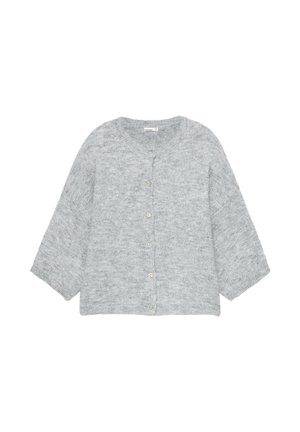 Jeune femme aux cheveux attachés portant un cardigan gris boutonné et un jean large bleu clair, debout dans une rue pavée.