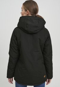 Giacca nera isolata con cappuccio, caratterizzata da una texture liscia, un retro leggermente allungato e un piccolo etichetta in pelle vicino all'orlo.