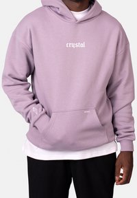 Sweatshirt à capuche lavande avec poche avant, comportant le texte "crystal" en blanc et un petit accent graphique sur la manche. Tissu doux et texturé.
