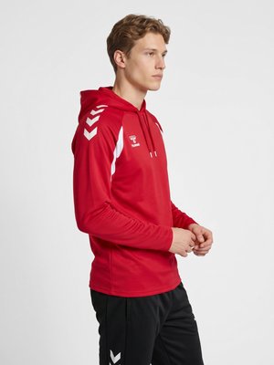 Giovane uomo con felpa sportiva rossa con cappuccio, strisce bianche a chevron sulle maniche e pantaloni neri, in piedi davanti a uno sfondo bianco.