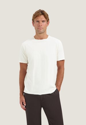 Man with short light brown hair wearing a plain white short-sleeve t-shirt and dark pants against a light gray background.