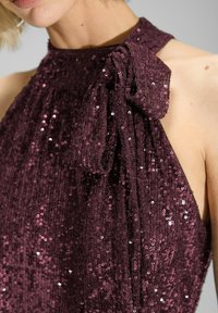 Abito senza maniche color bordeaux con paillettes; presenta un'acca di fiocco sul colletto. Texture liscia; riflette la luce con dettagli scintillanti.