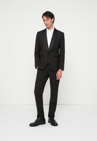 Jeune homme portant un smoking noir avec revers en satin, une chemise blanche, des chaussures de soirée noires, se tenant contre un fond blanc uni.