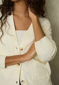 Cardigan avorio realizzato in morbido tessuto, con scollatura a v, maniche a coste, due tasche frontali e bottoni rotondi lungo il davanti.