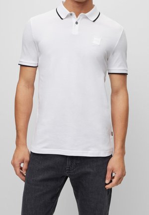 Polo blanc avec un col et des bordures de manches noirs, présentant un petit logo et deux boutons au cou ; fabriqué dans un tissu lisse.