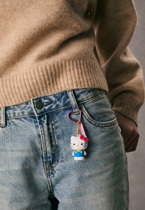 Kľúčový prsteň Hello Kitty pripevnený na svetlomodrom denimovom vrecku, s ružovým srdiečkovým uzáverom. Kľúčový prsteň zobrazuje kreslenú postavičku v modrých šatách.