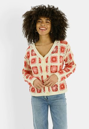 Mujer con cabello rizado sonriendo, vestida con un cárdigan de crochet crema y rojo y jeans azules, posando contra un fondo blanco.