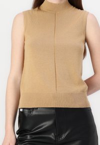 Jasnobrązowy bezrękawnik z dzianiny z półgolfem, drobną prążkowaną teksturą i pionowymi szwami. Połączony z czarnymi lśniącymi skórzanymi spodniami.