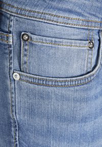 Gros plan sur un jean en denim bleu clair montrant la poche avant détaillée avec rivets métalliques et coutures jaunes.