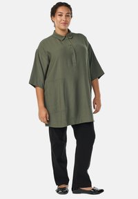 Frau steht da und trägt ein übergroßes, olivgrünes Hemd mit Vorderentaschen, schwarze Hose und schwarze Lacklederschuhe mit Schleifendetails.