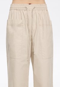 Beige lightweight pants with an elastic waistband, drawstring, and two large front pockets. Smooth texture with visible stitching details.