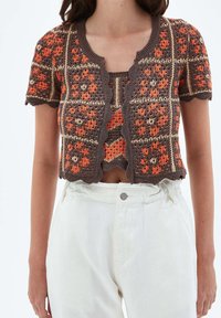 Mujer vistiendo un cárdigan corto de manga corta de ganchillo en marrón y naranja con patrones florales, combinado con pantalones blancos de cintura alta.