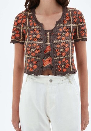 Mujer vistiendo un cárdigan corto de manga corta de ganchillo en marrón y naranja con patrones florales, combinado con pantalones blancos de cintura alta.