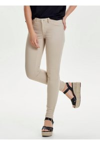 Mujer vestida con pantalones beige ajustados y sandalias negras de cuña con correas en el tobillo, de pie con una pierna ligeramente levantada.