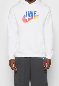 Person som bär en vit Nike-huvtröja med blå, röd och gul logotyp samt mörkgrå shorts med flerfärgade Nike-logotyper.
