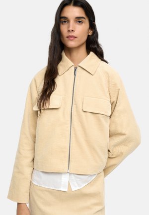 Kobieta o długich ciemnych włosach, ubrana w beżową kurtkę sztruksową na zamek z klapami na kieszeniach, na białej koszuli.